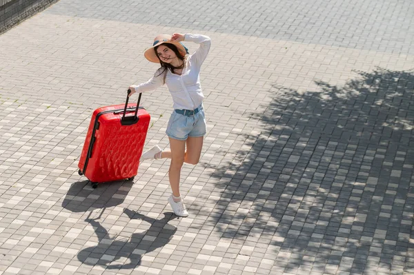 Glückliche kaukasische junge Frau mit Hut und kurzen Hosen, die in offenem Alter einen großen roten Koffer hält — Stockfoto