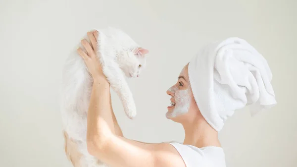 Kobieta z ręcznikiem na włosach i glinianą maską na twarzy trzyma białego puszystego kota na białym tle. — Zdjęcie stockowe