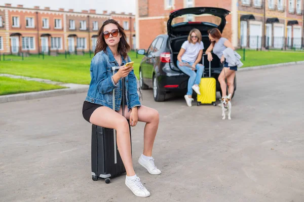 Drei Freundinnen gehen auf Roadtrip. Junge Frau posiert mit einem Koffer. Zwei Mädchen sitzen auf dem Kofferraum eines Autos und betrachten eine Landkarte. — Stockfoto