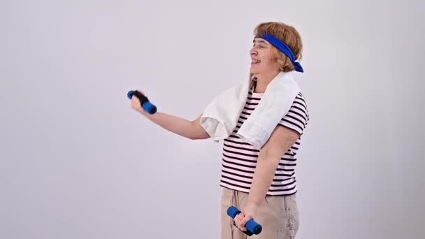 Uma mulher idosa com uma bandagem azul na cabeça treina com halteres em um fundo branco — Vídeo de Stock