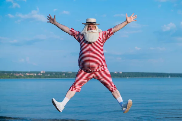Un uomo anziano in costume da bagno retrò a righe e Boater corre e salta lungo la spiaggia. Un vecchio dai capelli grigi allegro con la barba in un cappello riposa al mare. — Foto Stock