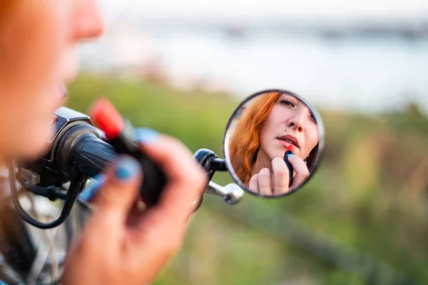 Eine Frau auf einem Motorrad blickt in den Spiegel und trägt Lippenstift auf. Spiegelbild eines rothaarigen Bikermädchens. — Stockfoto