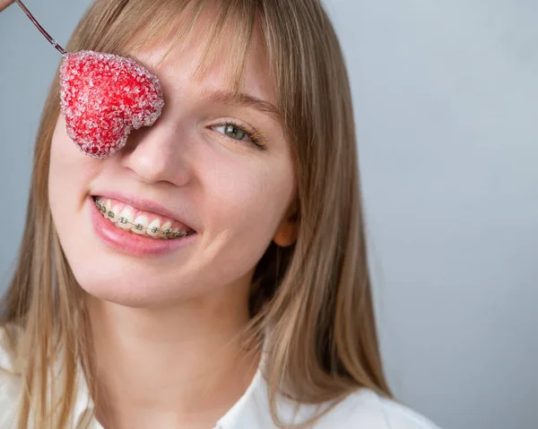 Söt kvinna med hängslen på tänderna håller ett godis i form av ett hjärta på vit bakgrund — Stockfoto