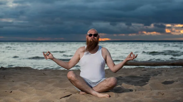 Лысый мужчина с рыжей бородой практикует йогу на пляже на закате. Забавный чувак в футболке и солнечных очках медитирует на берегу моря. — стоковое фото