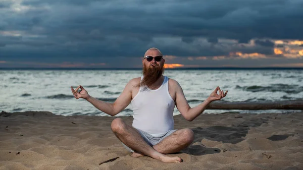 Un uomo calvo con la barba rossa pratica yoga sulla spiaggia al tramonto. Un tizio divertente con una maglietta e occhiali da sole medita sulla riva del mare. — Foto Stock