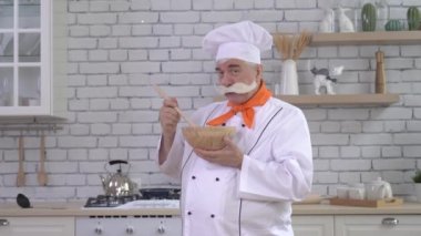 Dost canlısı yaşlı bir şef mutfakta bir tabak tutuyor. Gri saçlı, kalın gri bıyıklı bir adam mutfakta çalışıyor.