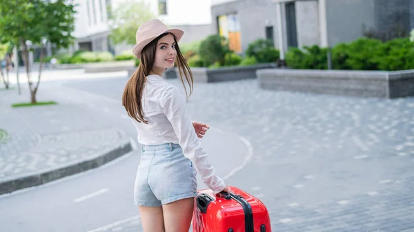 Junge Kaukasierin mit Hut und kurzer Hose steht mit rotem Koffer auf der Straße. — Stockfoto