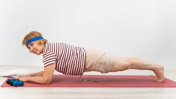 A happy old woman doing a plank on a sports mat. Elderly lady doing fitness exercises on white background. — Stock Photo, Image