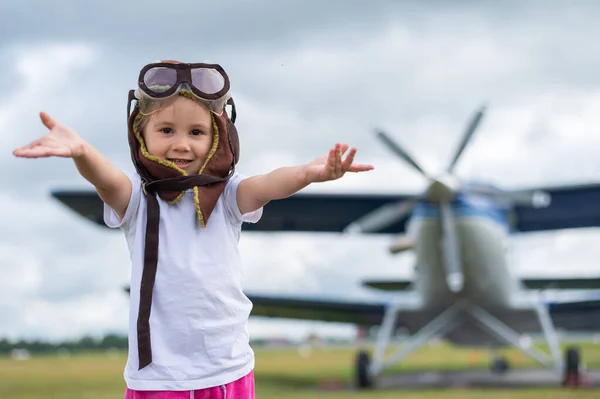 Una bambina carina vestita con un cappello e occhiali di un pilota sullo sfondo di un aeroplano. Il bambino sogna di diventare un pilota. — Foto Stock