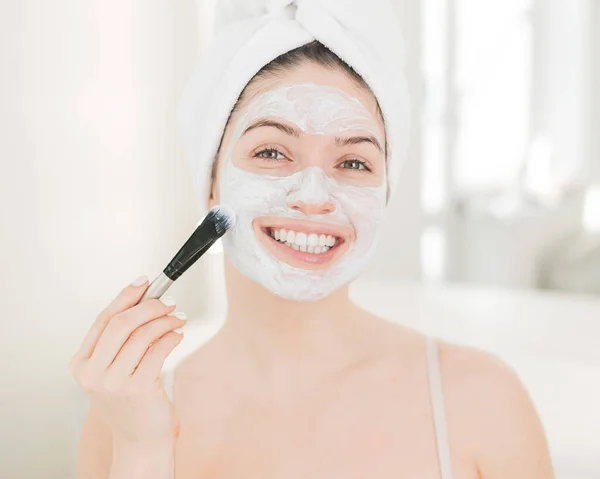 Mulher branca bonita com uma toalha em seu cabelo aplica uma máscara de argila. Cuidar da beleza em casa — Fotografia de Stock