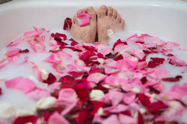 Funny picture of a man taking a relaxing bath. Close-up of male feet in a bath with foam and rose petals — Stock Photo, Image