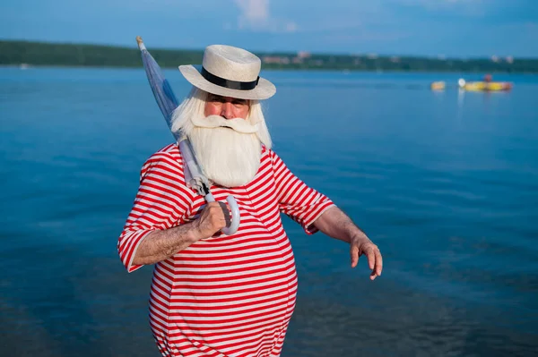 Un anziano in costume da bagno classico cammina lungo la spiaggia con un ombrellone in una calda giornata estiva — Foto Stock