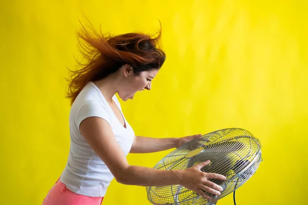 Krásná rusovlasá žena stojí nad velkým elektrickým ventilátorem na žlutém pozadí. Dívka s vlasy rozvíjejícími se ve větru. Zařízení pro chlazení vzduchu. — Stock fotografie