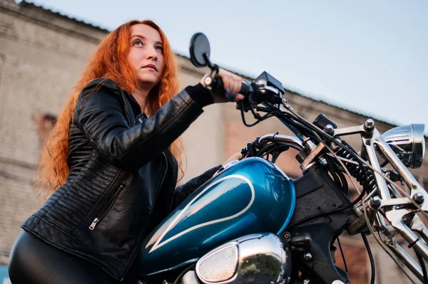 Lockige rothaarige Frau in schwarzer Lederjacke sitzt auf einem Motorrad. Porträt eines ernsthaften Mädchens, das Fahrrad fährt. — Stockfoto