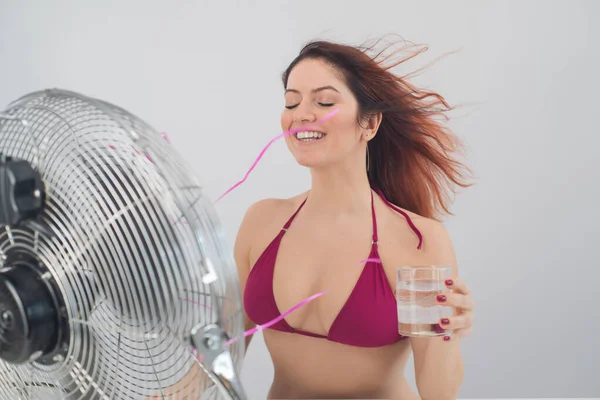 Κοκκινομάλλα χαμογελαστή γυναίκα σε ένα μπικίνι πίνει ένα κρύο ποτό και απολαμβάνει τον άνεμο που φυσάει από έναν ηλεκτρικό ανεμιστήρα σε λευκό φόντο. τον έλεγχο του κλίματος σε μια ζεστή μέρα του καλοκαιριού — Φωτογραφία Αρχείου