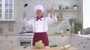 Dost canlısı, yaşlı bir şef aletlerini mutfakta kondüktör gibi sallıyor. Adam bir kepçe tutuyor ve çırpıyor.