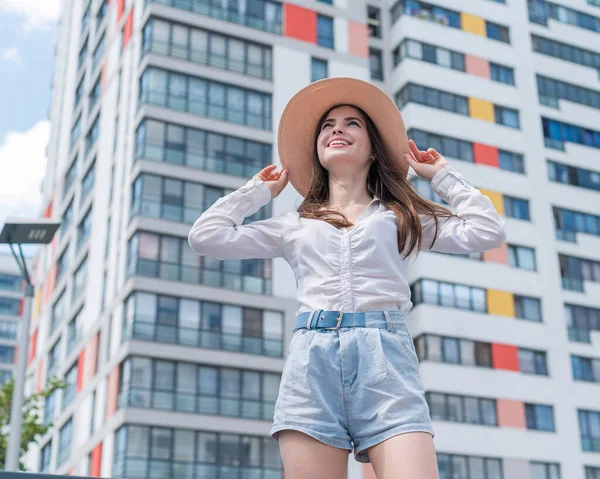 Feliz mujer joven caucásica en un sombrero y pantalones cortos camina en una calle de la ciudad en un día caluroso de verano —  Fotos de Stock