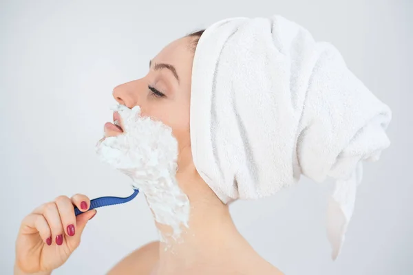 Une femme avec une serviette éponge blanche sur la tête et avec de la mousse à raser sur le visage se rase sur un fond blanc — Photo