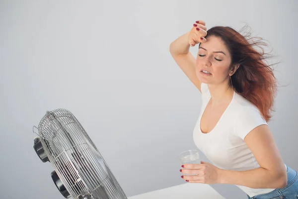 Una mujer caucásica pelirroja se enfría junto al ventilador eléctrico y bebe una bebida fría. Climatización en el apartamento — Foto de Stock