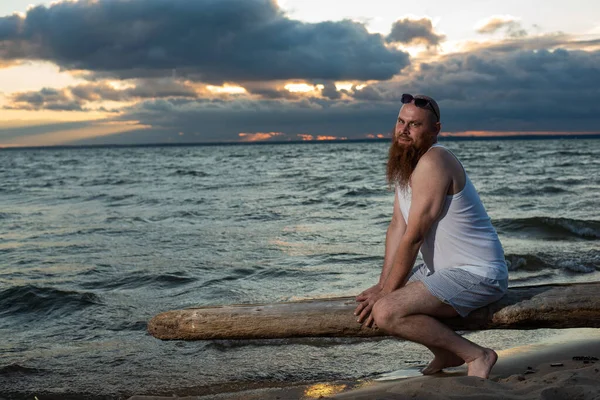 Funny bald man with red beard posing on the beach at sunset. A humorous male parody of a glamorous girl.