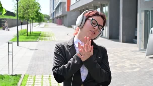 Mujer de negocios caucásica en un traje escuchando música con auriculares y cantando junto a bailar al aire libre — Vídeo de stock