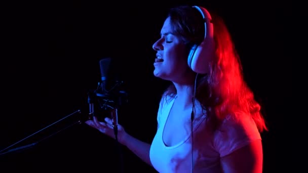 Caucasian woman in headphones singing into a microphone in neon light on a black background. An emotional girl is recording a song in a recording studio — Stock Video