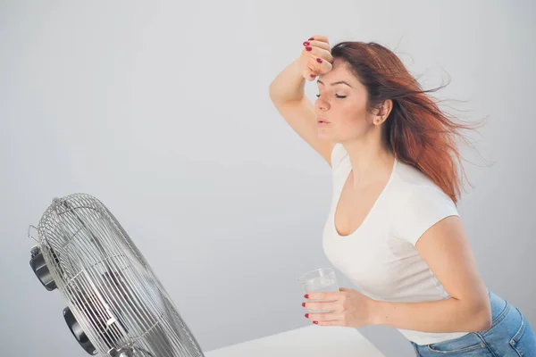 Punatukkainen valkoihoinen nainen vilustuu tuulettimen vieressä ja juo kylmää juomaa. Ilmastonsäätö asunnossa — kuvapankkivalokuva