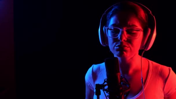 Caucasian woman in glasses and headphones sings into a microphone in neon light on a black background. An emotional girl is recording a song in a recording studio — Stock Video