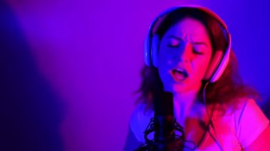 Kulaklıklı beyaz kadın, neon ışıkta mikrofona şarkı söylüyor. Duygusal bir kız kayıt stüdyosunda şarkı kaydediyor.