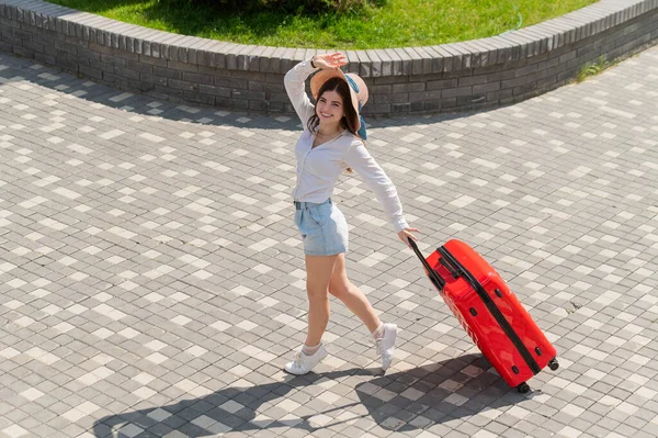 Glückliche kaukasische junge Frau mit Hut und kurzen Hosen, die in offenem Alter einen großen roten Koffer hält — Stockfoto