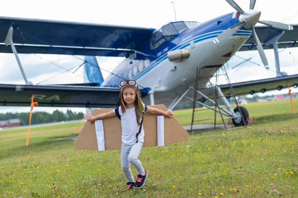 Een klein meisje in een pilotenkostuum met kartonnen vleugels loopt op het gazon tegen de achtergrond van het vliegtuig. Een kind met een hoed en een bril droomt van vliegen in een vliegtuig. — Stockfoto