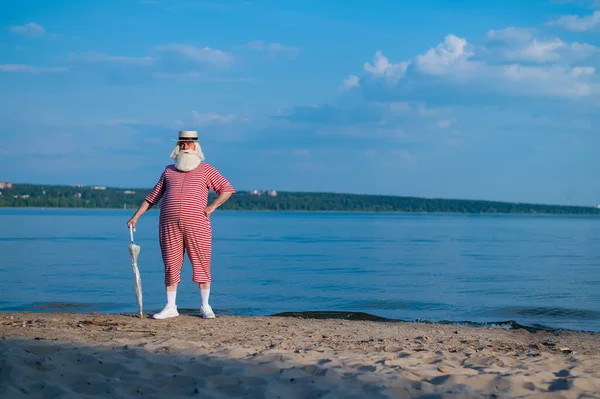 Пожилой человек в классическом купальнике прогуливается по пляжу с зонтиком в жаркий летний день — стоковое фото