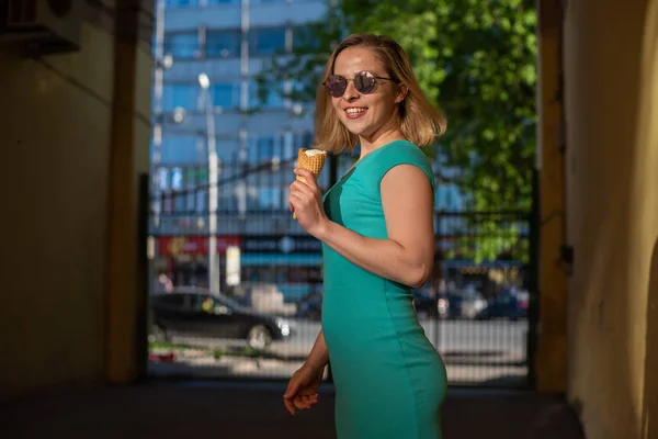 Eine glückliche Frau in einem türkisfarbenen Kleid steht in einem gewölbten Gang und isst an einem warmen Sommertag einen Waffelkegel. Die schöne Blondine mit Sonnenbrille genießt Eis beim Gehen. Sommer-Dessert. — Stockfoto