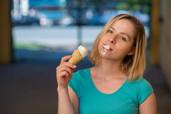 Porträtt av en söt tjej i grön klänning går ut och äter dessert. Vacker blondin njuter av en kon med glass en vacker sommardag. Livet är ett nöje — Stockfoto