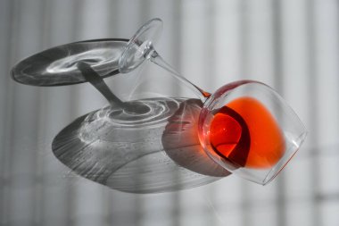 Bir kadeh kırmızı şarap yan yatar beyaz bir masada, panjurların gölgesinde.