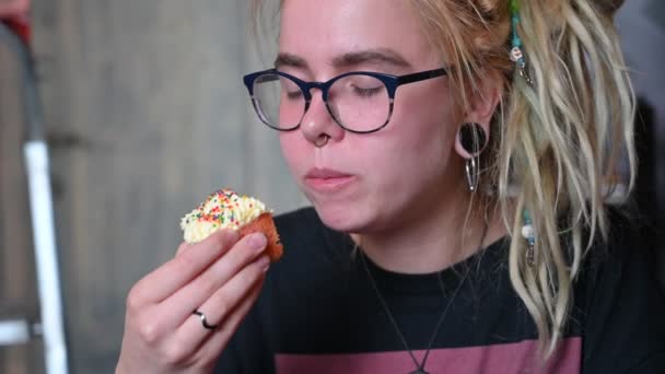 An unusual girl with dreadlocks on her head and a piercing eats a cupcake in a cafe. A young woman in glasses is having breakfast with a cupcake against a gray wall. Eating habits. Insulin. — Stock Video