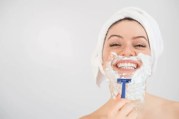 A woman with a white terry towel on her head and with shaving foam on her face shaves on a white background — Stock Photo, Image