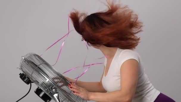 Red-haired Caucasian woman in sunglasses enjoys the breeze by the electric fan on a white background. Device cooling air. Slow motion — Stock Video