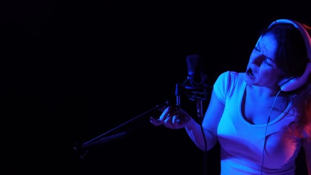 Mujer caucásica en auriculares cantando en un micrófono con luz de neón sobre un fondo negro. Una chica emocional está grabando una canción en un estudio de grabación — Vídeos de Stock