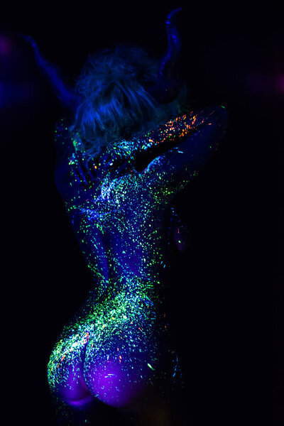 The back of a beautiful woman with horns on her head, the image of the devil. Woman in ultraviolet body painting. The girl is painted in fluorescent powder. Body art glowing in UV light..