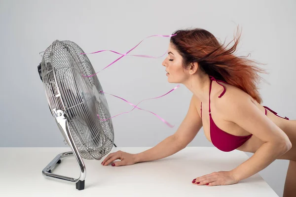 Bikinili kızıl saçlı beyaz kadın beyaz arka planda elektrikli vantilatörden soğuk hava akımını seviyor. Sıcak bir yaz gününde iklim kontrolü — Stok fotoğraf