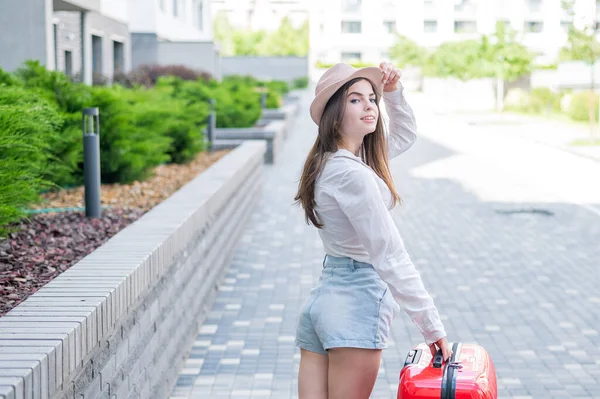 Jonge blanke vrouw in een hoed en korte broek staat op straat met een rode koffer. — Stockfoto