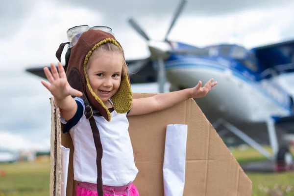 Ein nettes kleines Mädchen mit Mütze und Pilotenbrille auf dem Hintergrund eines Flugzeugs. Das Kind träumt davon, Pilot zu werden. — Stockfoto