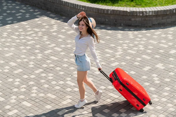 Glückliche kaukasische junge Frau mit Hut und kurzen Hosen, die in offenem Alter einen großen roten Koffer hält — Stockfoto