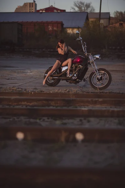 Mulher sexy bonita em terno de corpo de renda e saltos altos senta-se em uma motocicleta em um fundo industrial — Fotografia de Stock