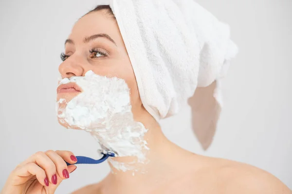 En kvinna med en vit frotté handduk på huvudet och med raklödder på ansiktet rakar sig på en vit bakgrund — Stockfoto