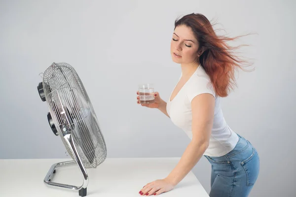 Punatukkainen valkoihoinen nainen vilustuu tuulettimen vieressä ja juo kylmää juomaa. Ilmastonsäätö asunnossa — kuvapankkivalokuva