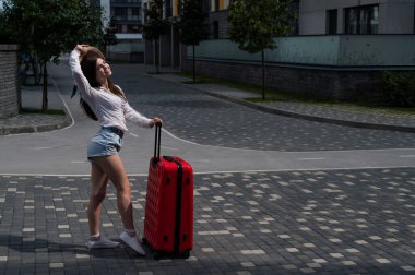 Kafkasyalı güzel esmer kadın, şapkalı, elinde bavuluyla şehir caddesinde poz veriyor..