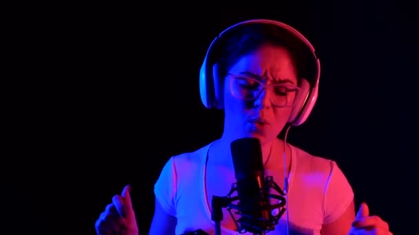 Fehér nő szemüvegben és fülhallgatóban neon fényben énekel egy mikrofonba, fekete háttérrel. Egy érzelmes lány felvesz egy dalt egy stúdióban. — Stock videók