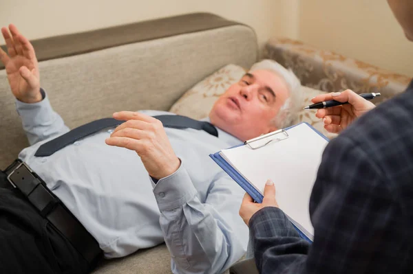 Um homem idoso se deita em um sofá durante uma consulta com um psicoterapeuta. Um idoso do sexo masculino em uma consulta individual com uma psicóloga do sexo feminino — Fotografia de Stock